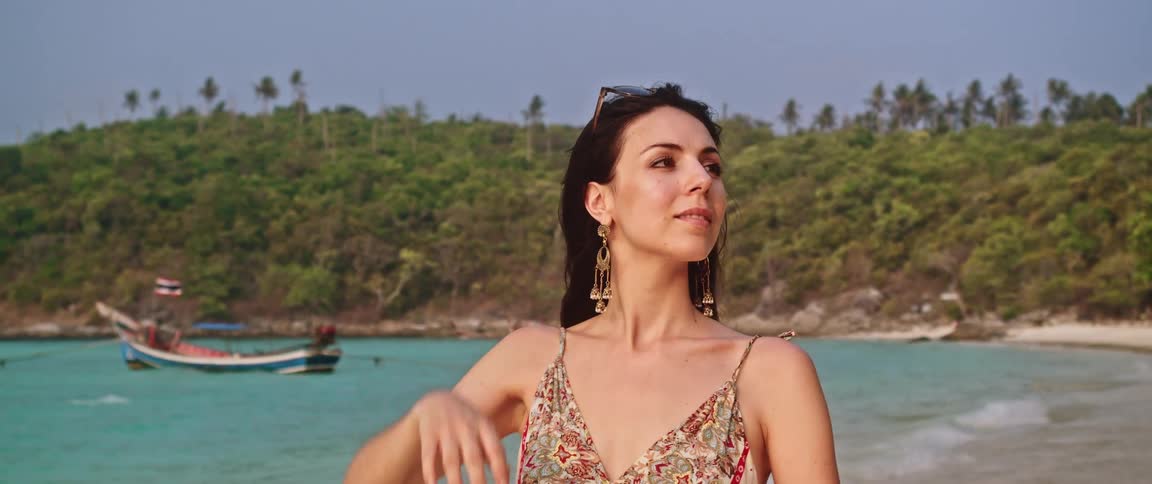 A thoughtful woman enjoys a serene moment on a tropical beach, looking out at the turquoise sea in slow motion