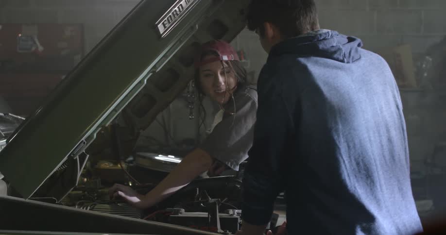 A woman works on a car in a garage, and a man interrupts her