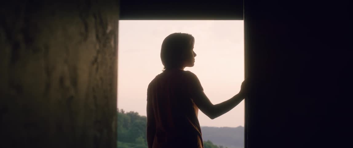 A woman walks toward a large window and gazes into the distance