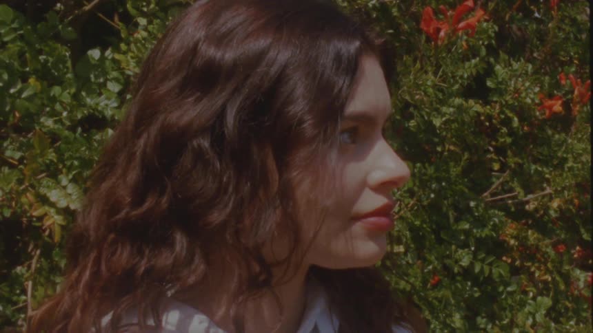 A beautiful young woman with long, wavy dark hair turns to look at the camera with a soft smile in a lush, sunlit garden