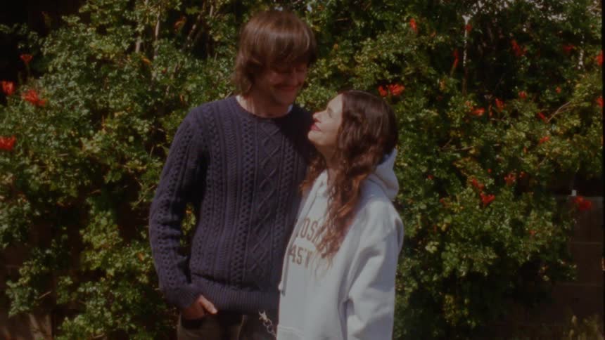 A man affectionately kisses his smiling girlfriend on the forehead while standing in a sunny garden