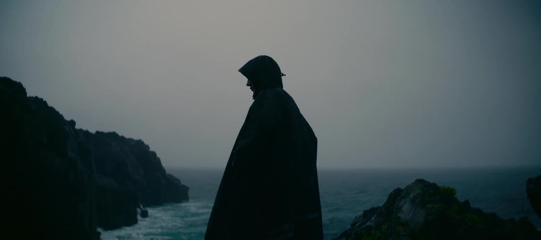 A silhouetted figure in a dark hooded cloak braves a storm on a rocky cliff overlooking the turbulent sea