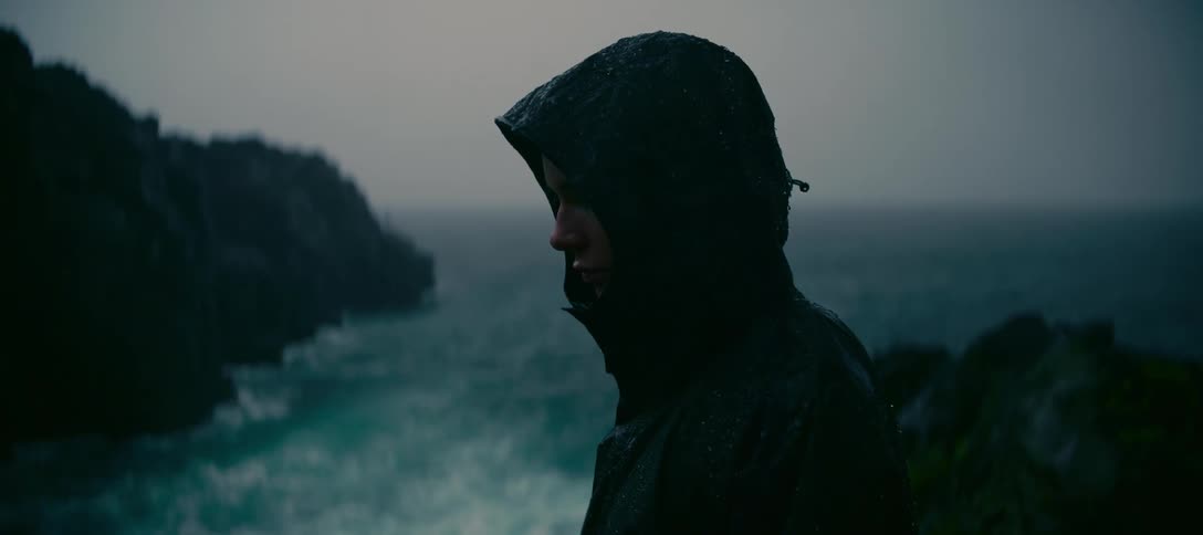 A cinematic shot of a sad man in a black hooded raincoat looking out over the stormy ocean on a gloomy day