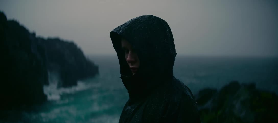 A mysterious figure in a dark hooded raincoat looks out at the stormy sea from a cliffside during a heavy downpour