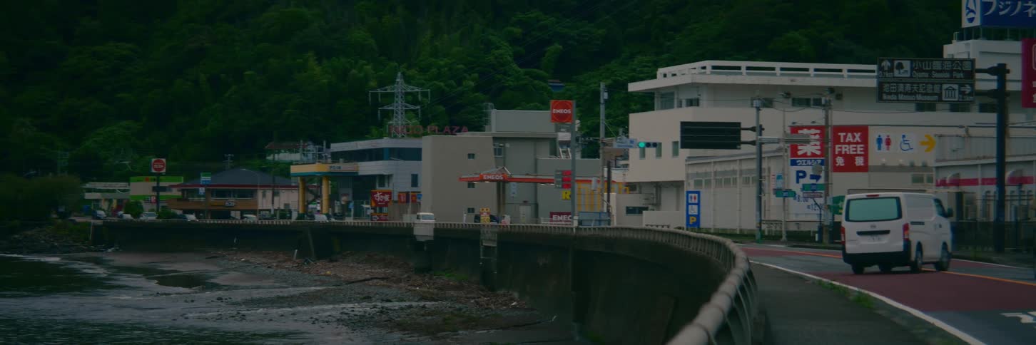 Cars drive along a quiet coastal road in a Japanese town on an overcast day