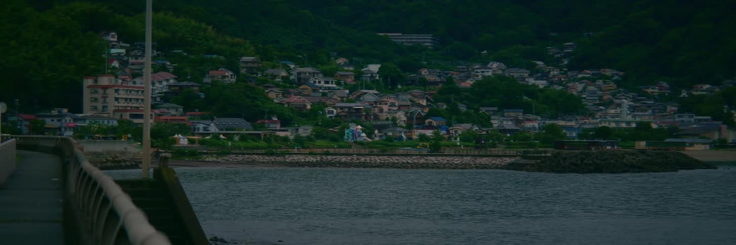 A serene view of a small Japanese coastal town nestled against a lush green hillside on an overcast day