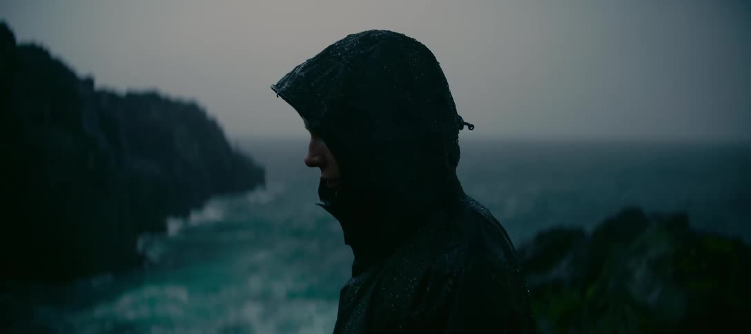 A lone figure in a hooded raincoat stands on a cliffside, looking down at the churning ocean in a moody downpour
