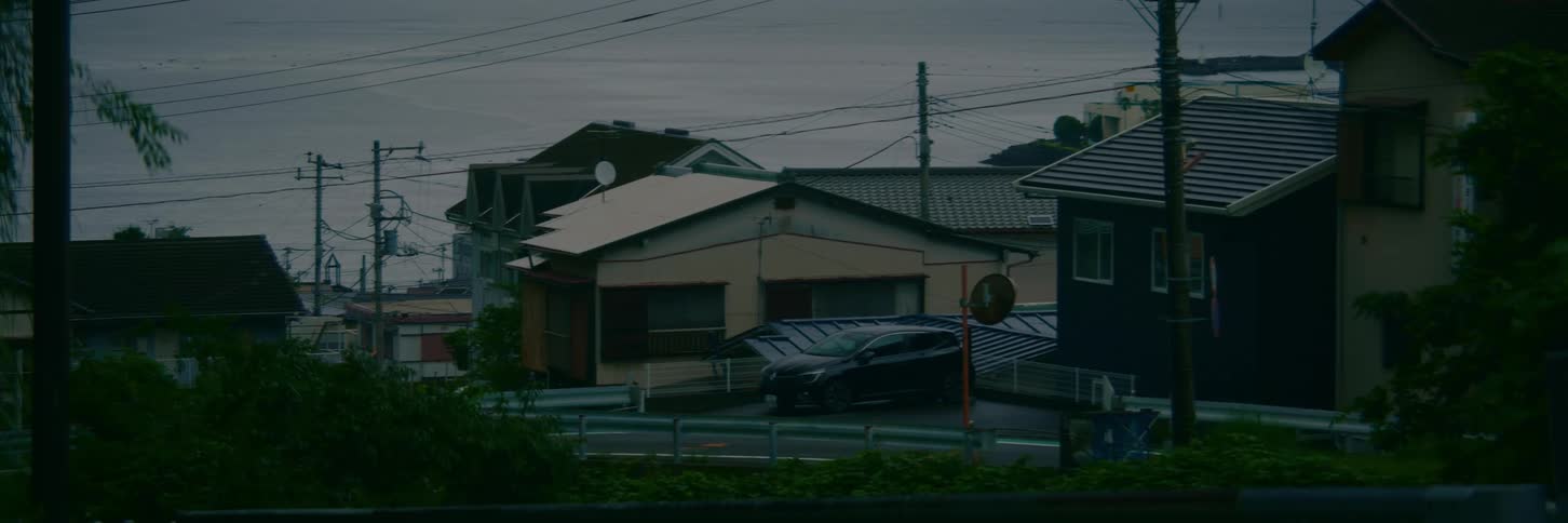 A static view of a quiet Japanese coastal town with traditional houses and a parked car under a gloomy, overcast sky