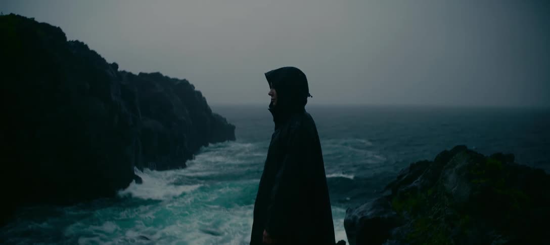A smiling person in a black rain poncho stands on a rocky cliff overlooking a turbulent sea during an overcast, stormy day