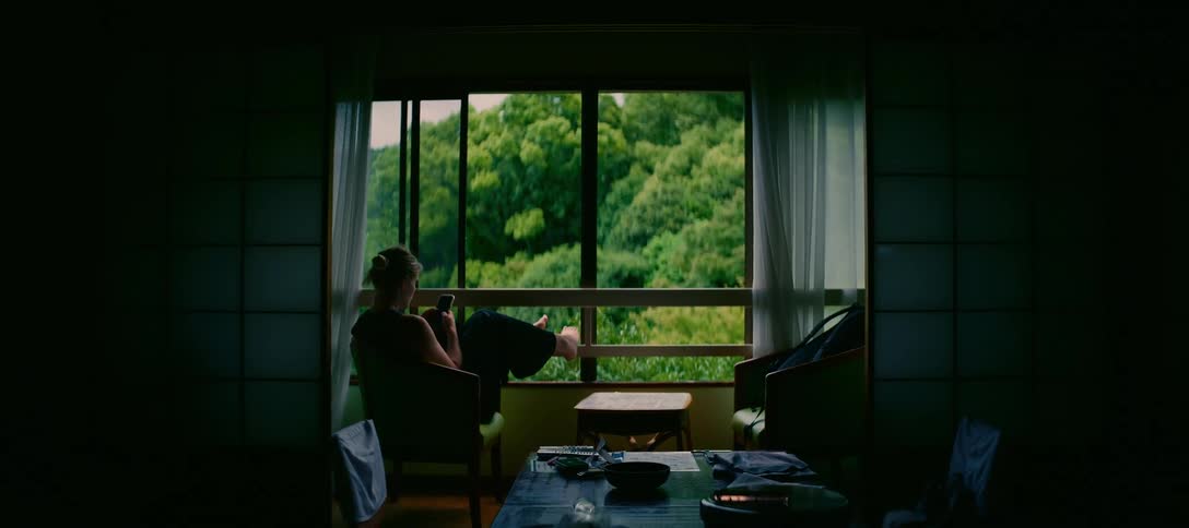 A woman relaxes on her phone in a traditional Japanese room overlooking a lush, green forest