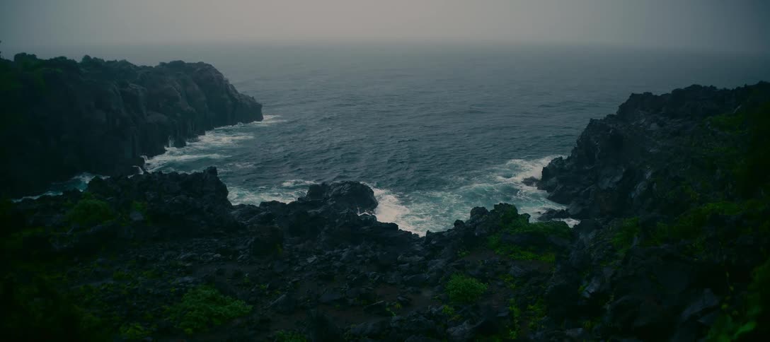Choppy ocean waves crash against a dark, volcanic rock coast on a moody and overcast day