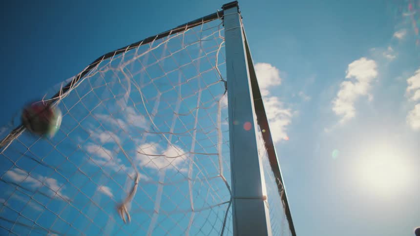 A low-angle, slow-motion shot captures a soccer ball hitting the back of the net against a bright blue sunny sky
