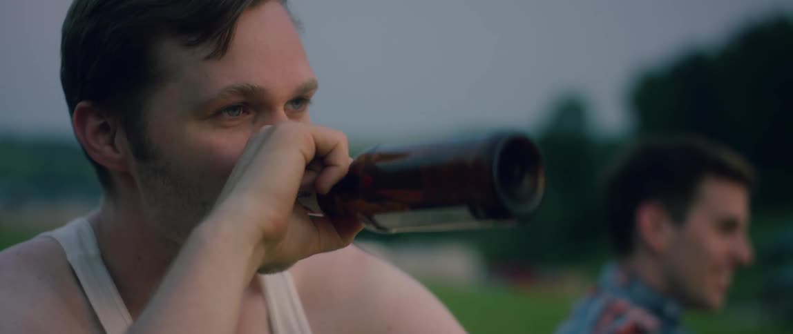 A man sitting outside laughs and drinks from a beer bottle