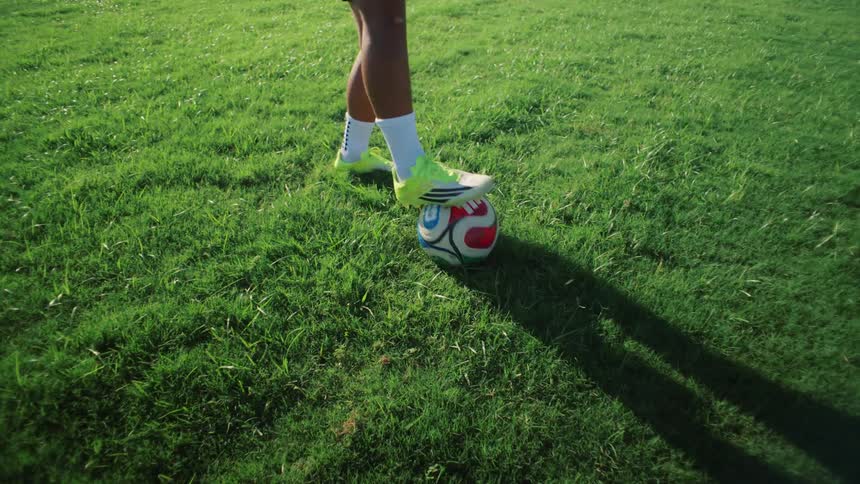 A soccer player's shadow follows a rolling ball across a grassy field before they stop it with their cleat