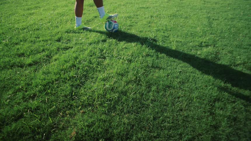 Dynamic POV shot follows a soccer player's shadow running to a ball on a green field, ending with their foot resting on top of it