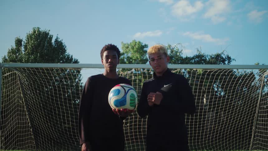Two serious young male soccer players pose for the camera with a ball in front of the goal on a sunny day