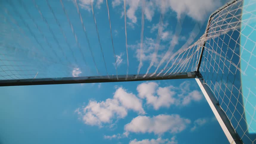 A slow-motion shot from inside a soccer goal shows a ball hitting the back of the net against a bright blue sky