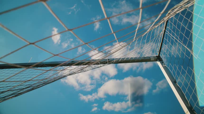 Low angle, slow motion shot of a soccer ball hitting the back of the net against a bright blue sky