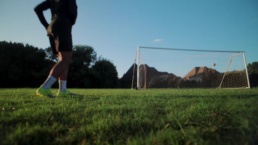 From a dramatic low angle, a soccer player kicks a ball and scores a goal in slow motion during a sunny evening