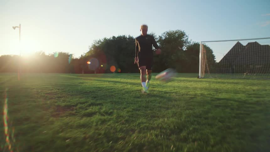 Low-angle tracking shot of a young man practicing soccer, kicking the ball on a grassy field at golden hour