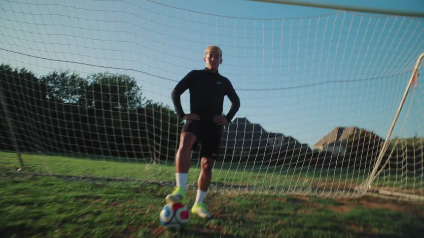 A confident young soccer player stands with his foot on the ball, posing in front of a goal on a sunny day as the camera pushes in