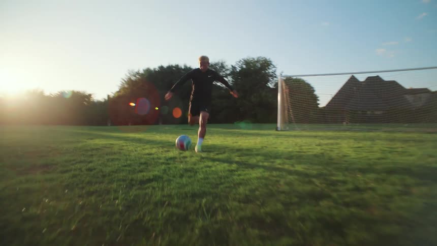 A young male soccer player dribbles a ball towards the camera on a green field during a sunny evening in slow motion