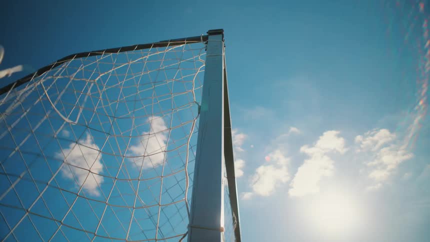 Low angle slow motion view of a soccer ball scoring a goal as it hits the net under a sunny blue sky