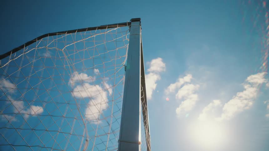A soccer ball scores a goal as it flies into the net in slow motion against a bright blue sky
