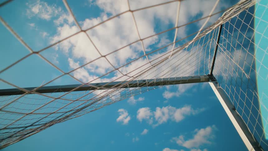 A soccer ball hits the back of the net from a low angle point of view, with a beautiful blue sky and clouds in the background