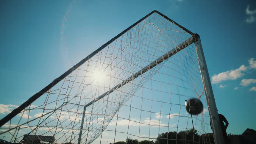 A soccer ball soars into the goal net in slow motion against a bright, sun-flared blue sky