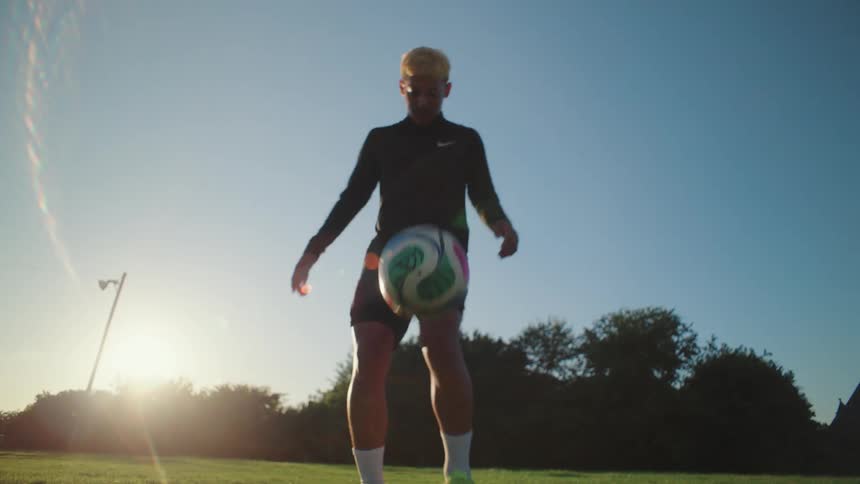 A skilled male soccer player with bleached hair juggles a ball in slow motion on a grassy field against the setting sun