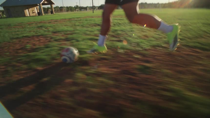 Dynamic point-of-view shot follows a soccer player dribbling a ball across a grassy field at sunset