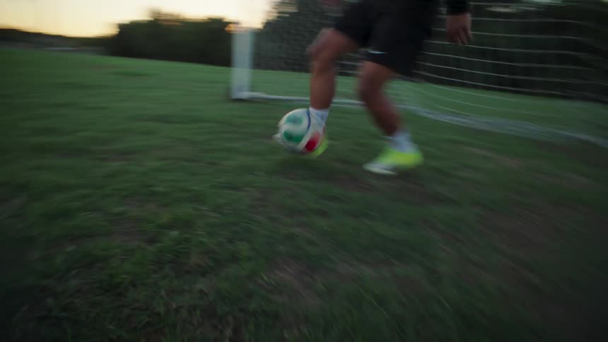Dynamic low-angle shot follows a soccer player's feet as they dribble and powerfully kick the ball on a grassy field at dusk