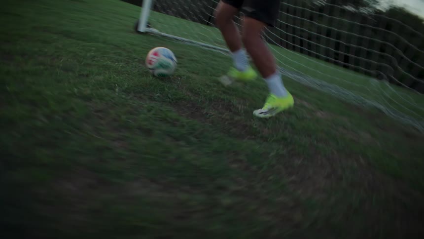 In slow motion, a soccer player dribbles a ball down the field and kicks it towards the goal at dusk