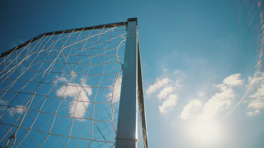 Low-angle slow-motion shot of a soccer ball hitting the back of the net on a sunny day with a blue sky