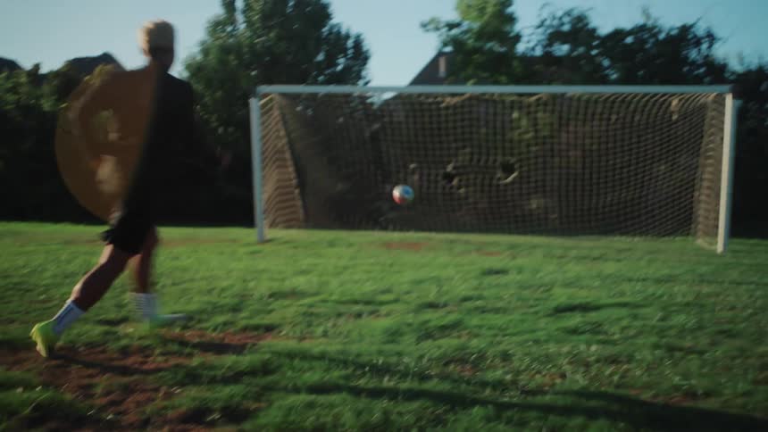 A soccer player dribbles a ball down a grassy field and kicks it into the goal in slow motion with cinematic lens flare