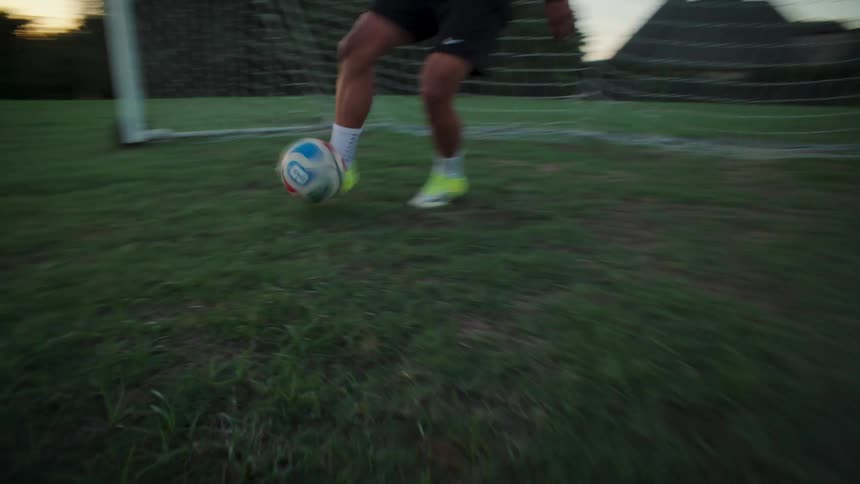 Low-angle view of a soccer player's feet dribbling a ball on a grassy field in front of a goal at dusk