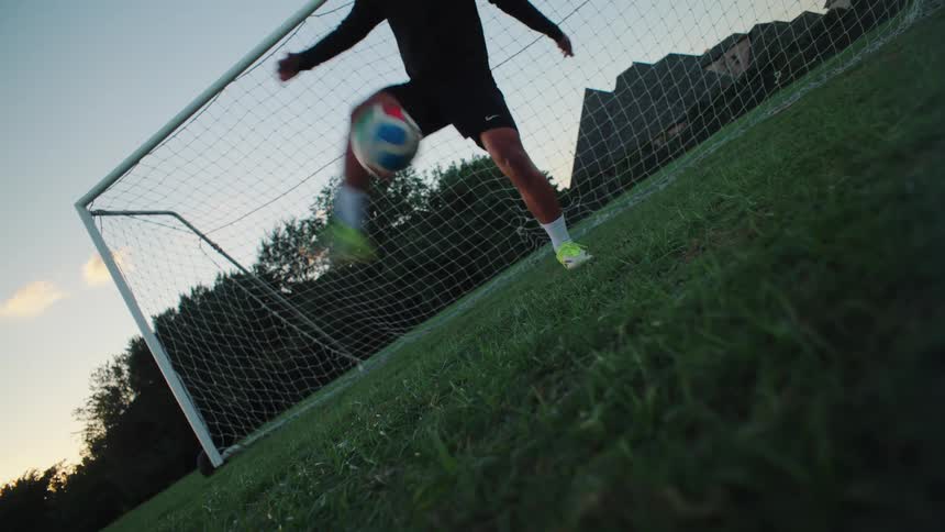 A skilled soccer player juggles a ball and performs a bicycle kick in slow motion in front of a goal at dusk