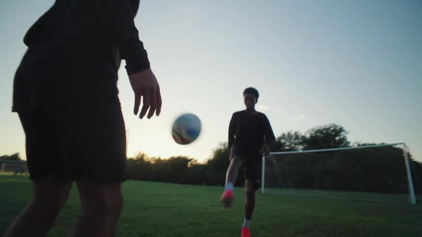 Dynamic slow motion reveals two young men skillfully juggling a soccer ball on a green field as the sun sets