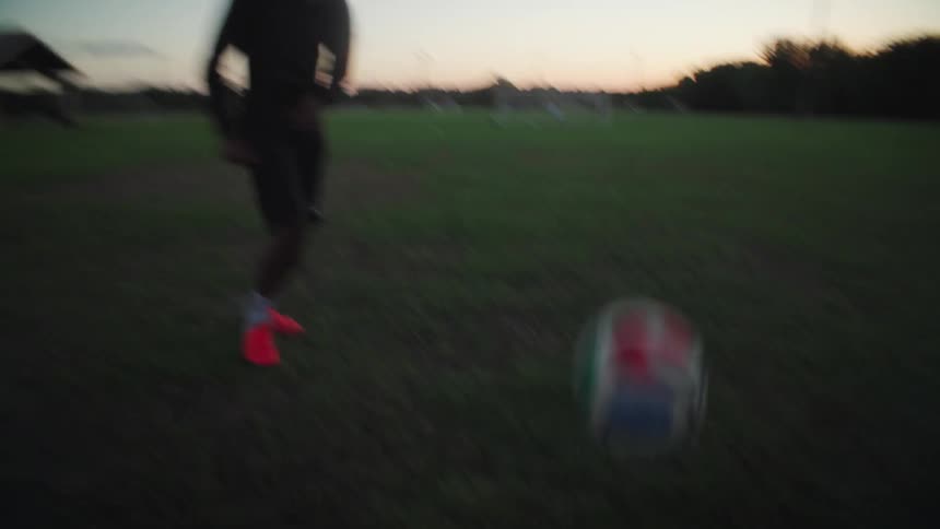 In slow motion, a young man juggles a soccer ball on a field at sunset before kicking it at the goal