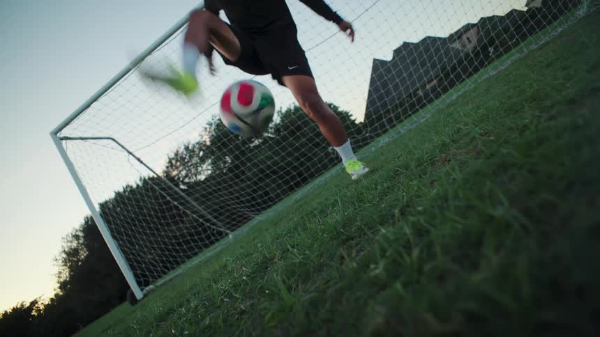 A low angle, slow motion shot of a soccer player juggling a ball before kicking it powerfully into the goal
