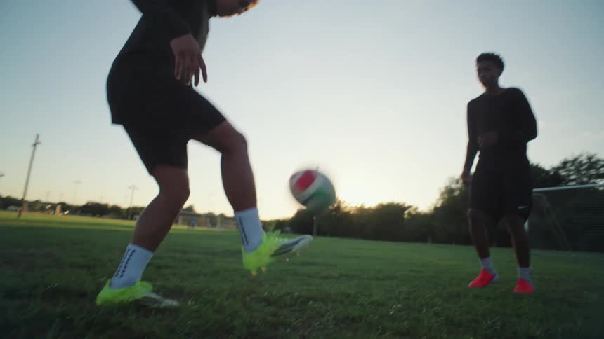 In a low-angle slow-motion shot, two athletic friends skillfully juggle a soccer ball on a grassy field during a beautiful sunset