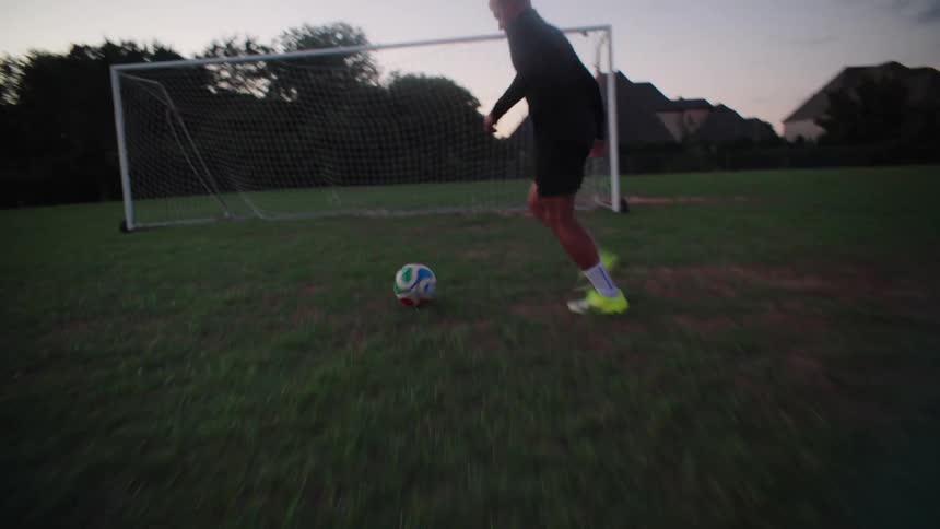 Dynamic low-angle shot follows a soccer player dribbling a ball down a grassy field and scoring a goal at dusk