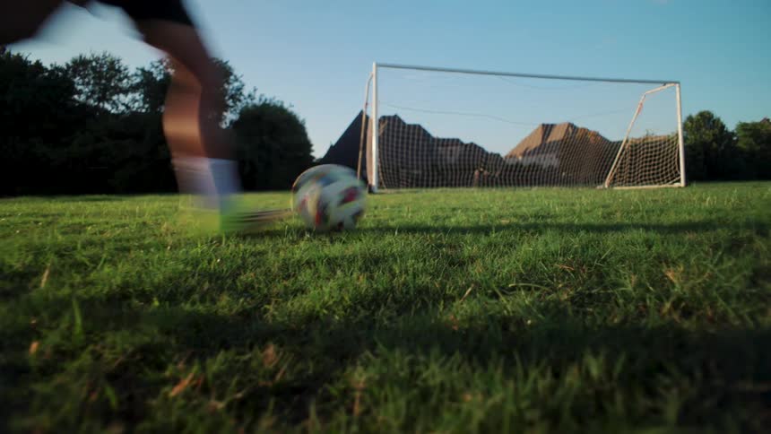 From a low angle, a soccer player kicks a ball into the goal while practicing on a lush green field at sunrise