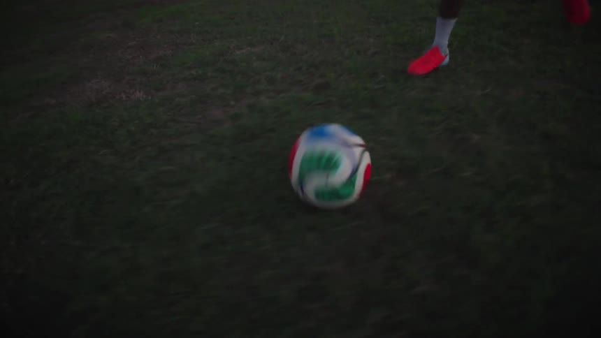 A male soccer player dribbles a ball down an empty field and kicks it towards the goal at dusk in slow motion
