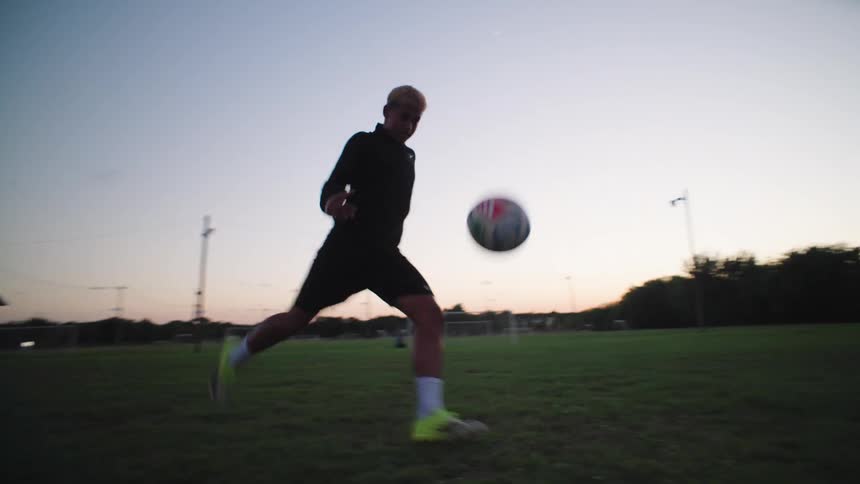A male soccer player practices juggling and kicking a ball towards the goal on a grassy field at dusk in slow motion