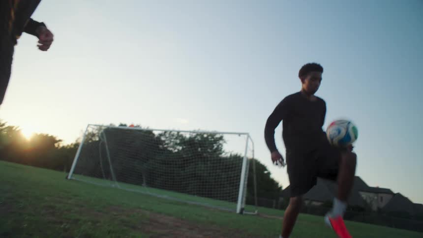 Two young athletic men showcase their soccer skills, juggling a ball back and forth in slow motion on a grassy field at sunset