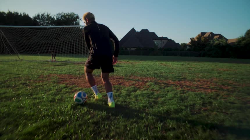 A soccer player in black athletic wear dribbles and juggles a ball down a grassy field toward the goal during a sunny morning practice