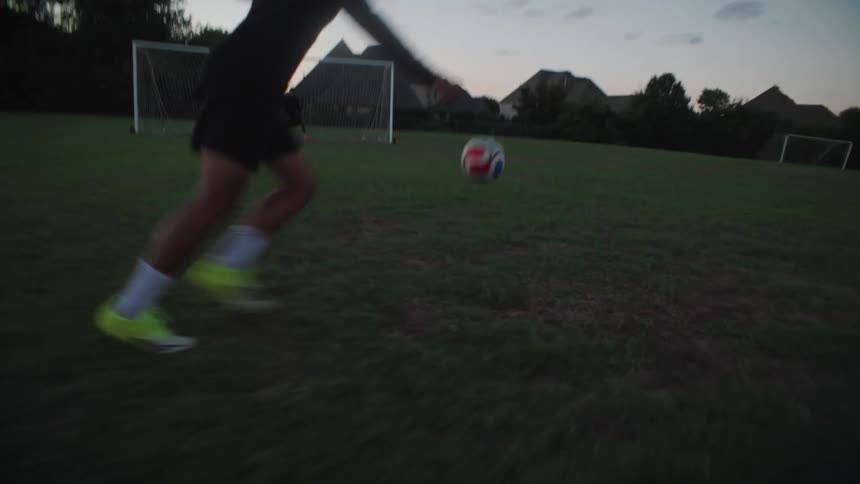 A soccer player controls a falling ball, dribbles, and kicks it towards the goal in a dynamic slow-motion shot at dusk