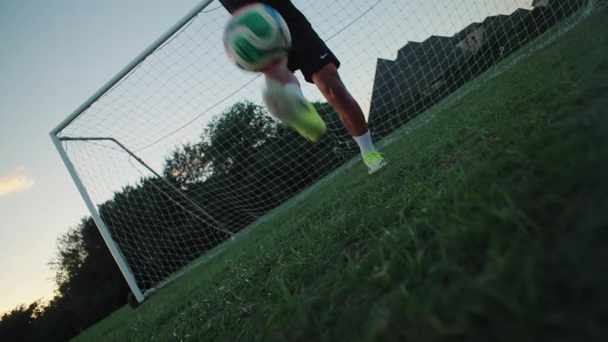 A soccer player juggles a ball in slow motion from a dramatic low angle in front of a goal at dusk
